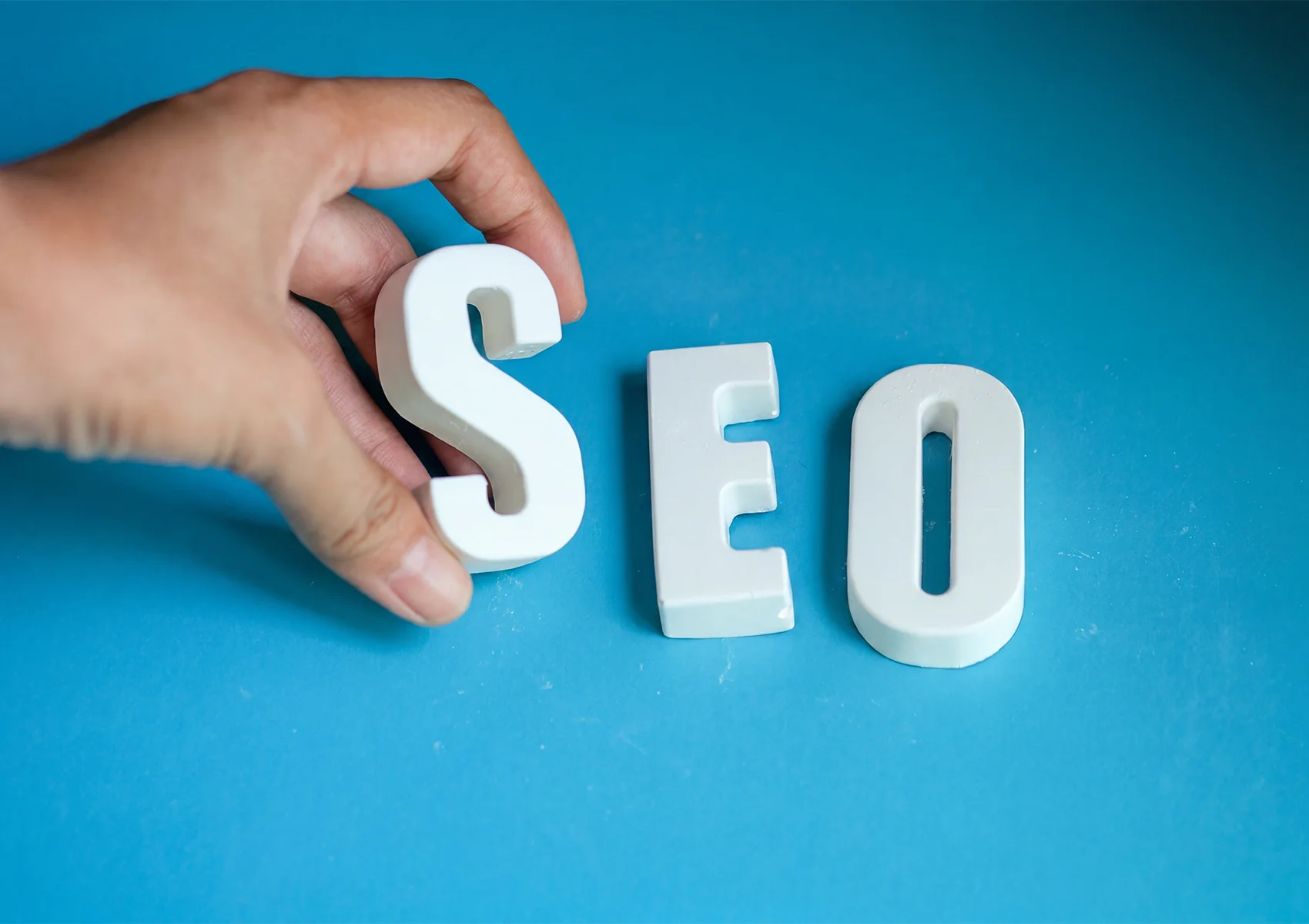 Hand arranging SEO letters on a blue background to illustrate the concept of search engine ranking and website visibility.